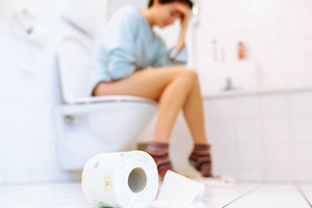Woman sitting uncomfortably on toilet with a toilet roll in foreground.